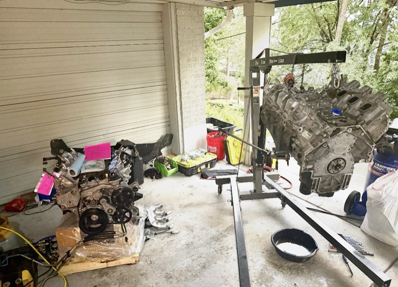 An engine hanging from an engine hoist in a carport during an engine swap, with a second engine on a pallet nearby.