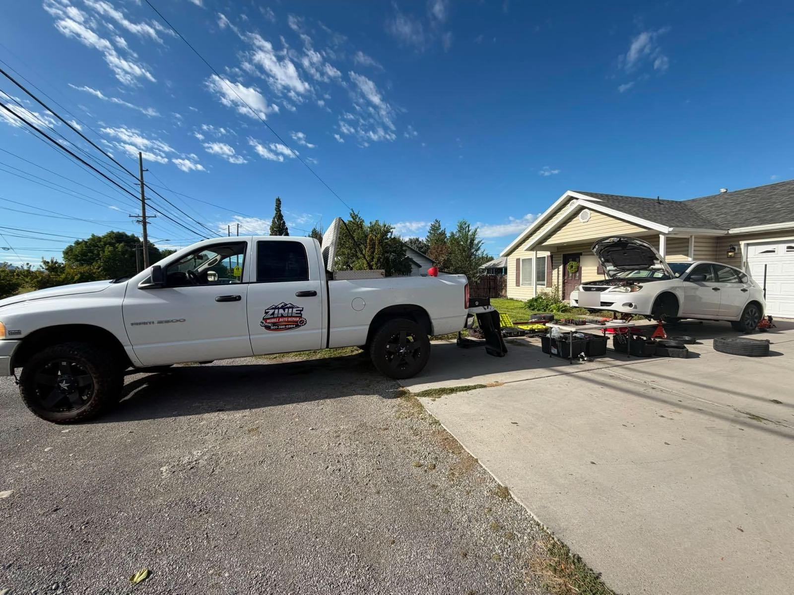 White work truck with a Zinie Mobile Auto Repair door decal parked in a driveway, with a car on a jack, its hood open and tools laid out on a table beside it.